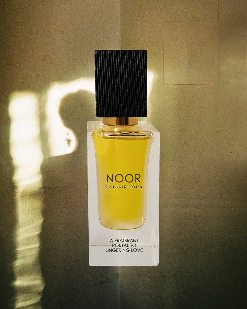 Perfume bottle labeled 'NOOR' on a textured wall with sunlight casting shadows.