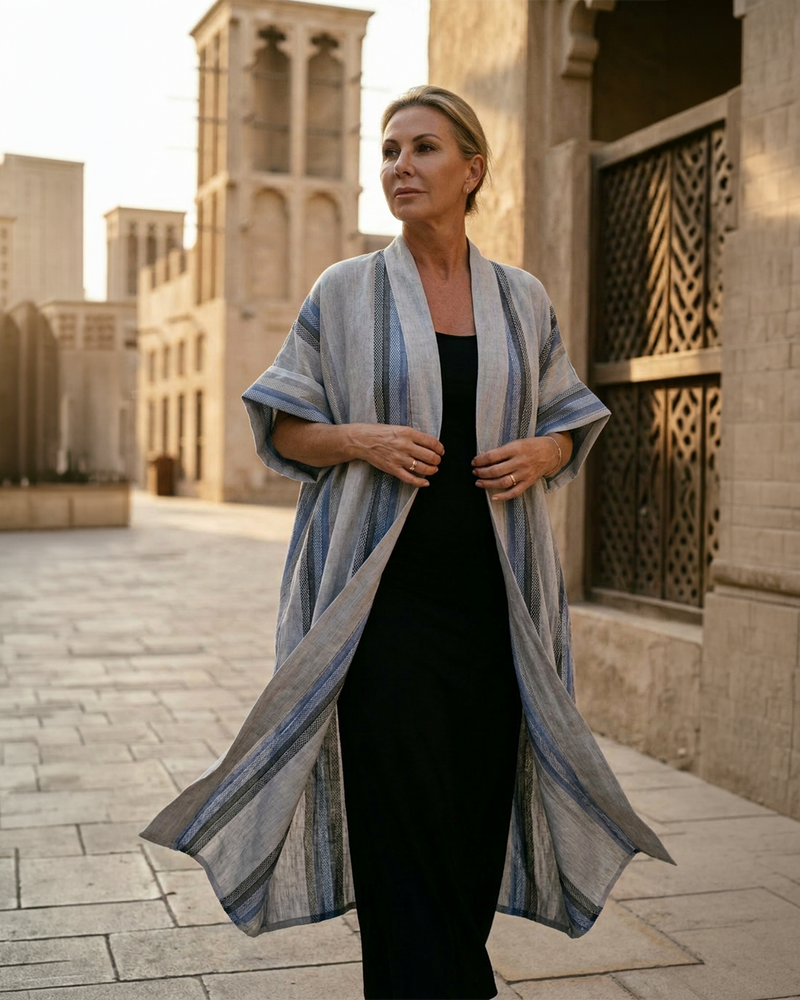 Natalie Noor wearing a long, light blue and gray striped robe over a long black fitted dress, standing in front of an architectural building. From the Sahara Resort Wear Collection