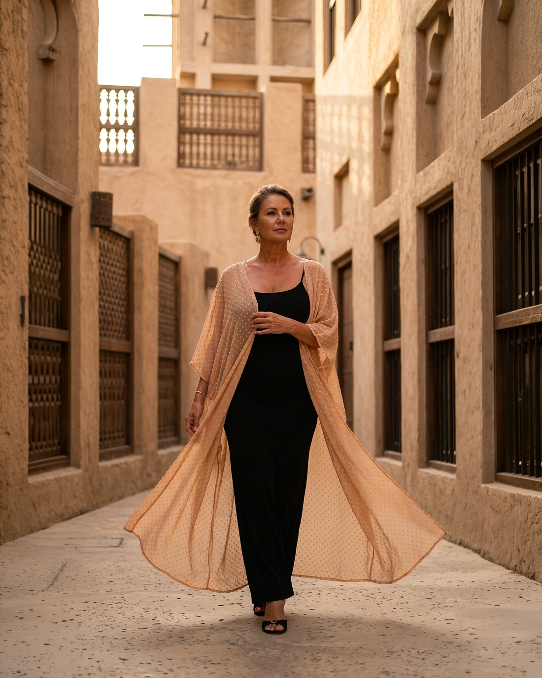 Natalie Noor in a black dress with a fluid nude uragiri chiffon robe, walking through an alleyway of old Dubai.