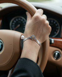 Hand with a silver cuff bracelet on a steering wheel inside a car. Sharafeh Collection by Natalie Noor - Jewelry lit by Desire