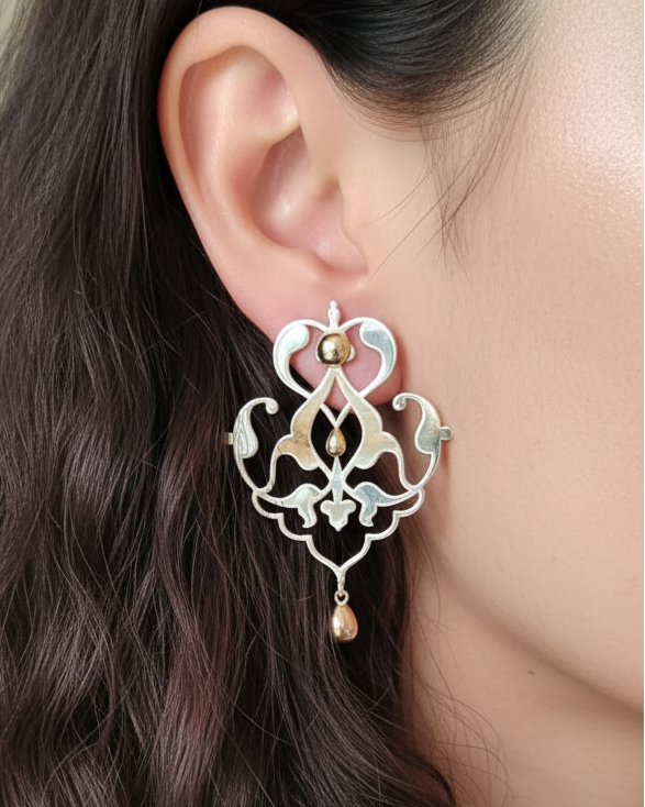 Close-up of a woman wearing an ornate earring in silver with golden inserts. Sharafeh collection, jewelry lit by desire, maison natalie noor