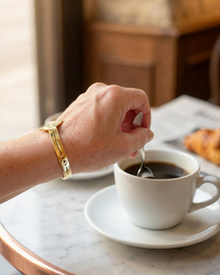 Person stirring a cup of coffee on a marble table with a croissant in the background. She is wearing a golden cuff bracelet form the Sharafeh Collection. Maison Natalie Noor, jewelry lit by Desire.