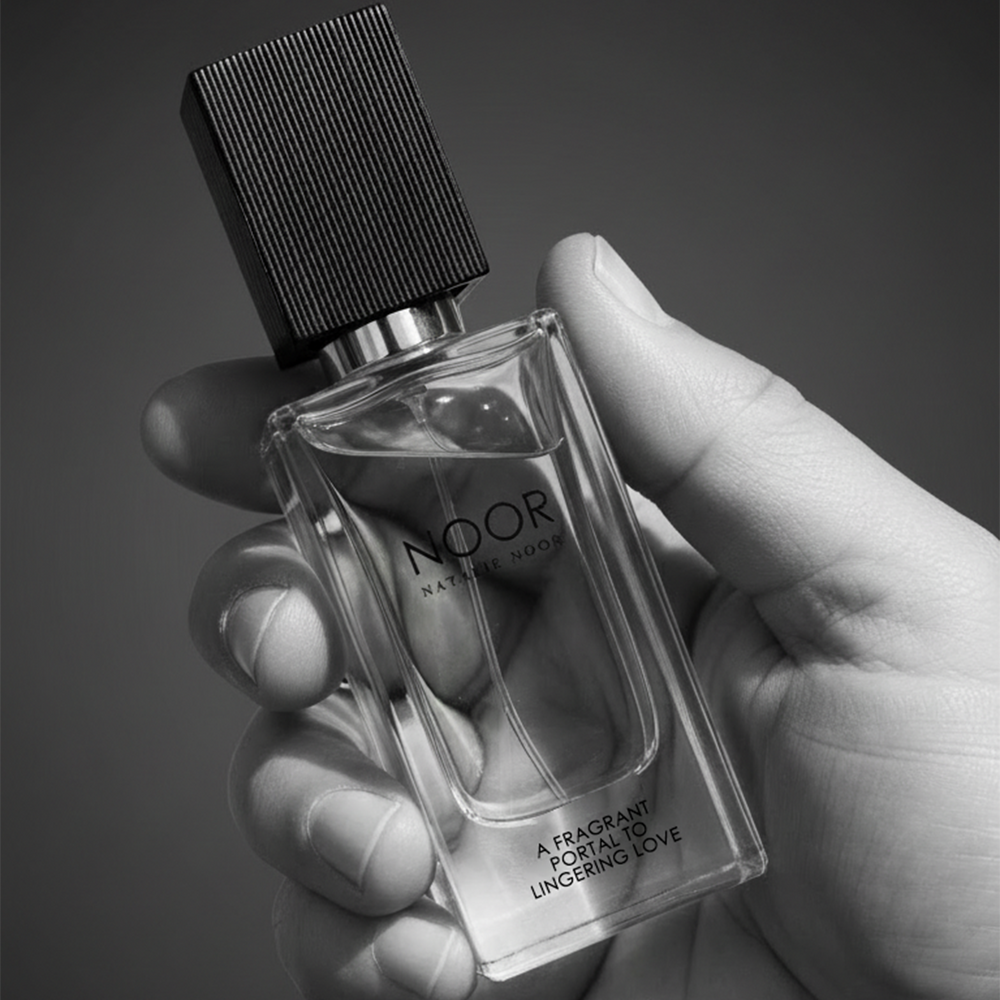 Hand holding a clear perfume bottle labeled 'NOOR' against a dark background. A Fragrant Portal to Lingering Love by Natalie Noor