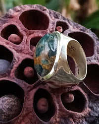 Silver ring with a green and golden apatite gemstone on a textured pink surface. Maison Natalie Noor, jewelry lit by desire.