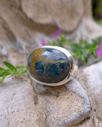 Silver signet ring with a blue and gold apattite cabochon gemstone on a stone surface with pink flowers. Maison Natalie Noor, jewelry lit by deisre.