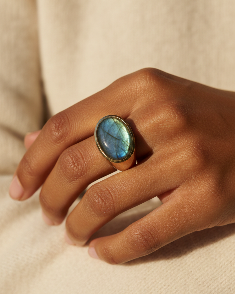 A woman's hand wearing a cabochon signet ring with a labradorite gemstone on a beige background. By Natalie Noor.
