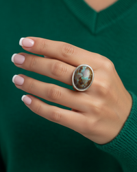 A woman's hand wearing a cabochon signet ring with a turquoise stone against a green jumper. By Natalie Noor