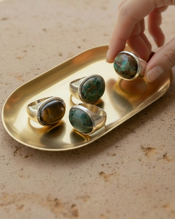 Sterling silver cabochon signet rings with gemstones on a golden tray, with a hand reaching for one of the rings. By Natalie Noor.