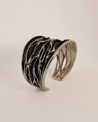 Silver cuff bracelet with wavy design on a light beige background. Plisse technique. Maison Natalie Noor, jewelry lit by desire.