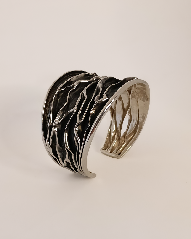Silver cuff bracelet with wavy design on a light beige background. Plisse technique. Maison Natalie Noor, jewelry lit by desire.