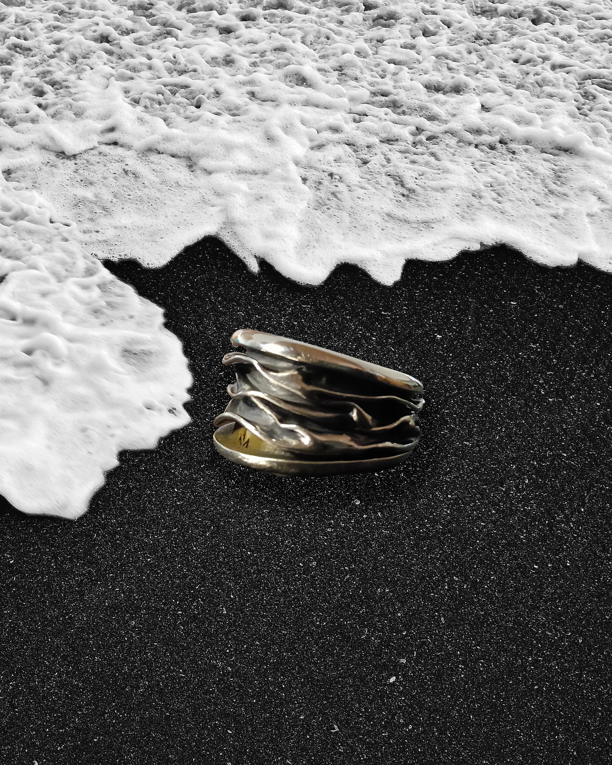 Silver ring on black sand with white foam at the water's edge. Maison Natalie Noor, jewelry lit by desire