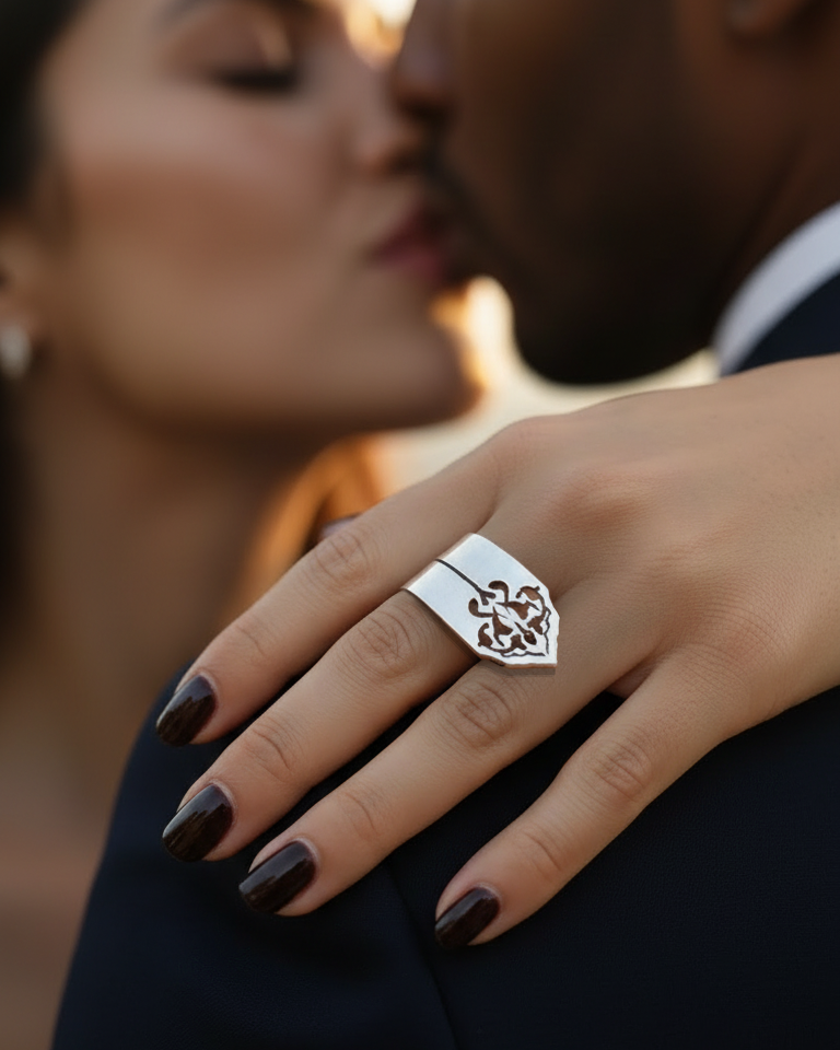 Close-up of a hand wearing a decorative ring with a blurred background of two people embracing. Silver plated ring from the Sharafeh Collection, Jewlery lit by Desire by Natalie Noor.
